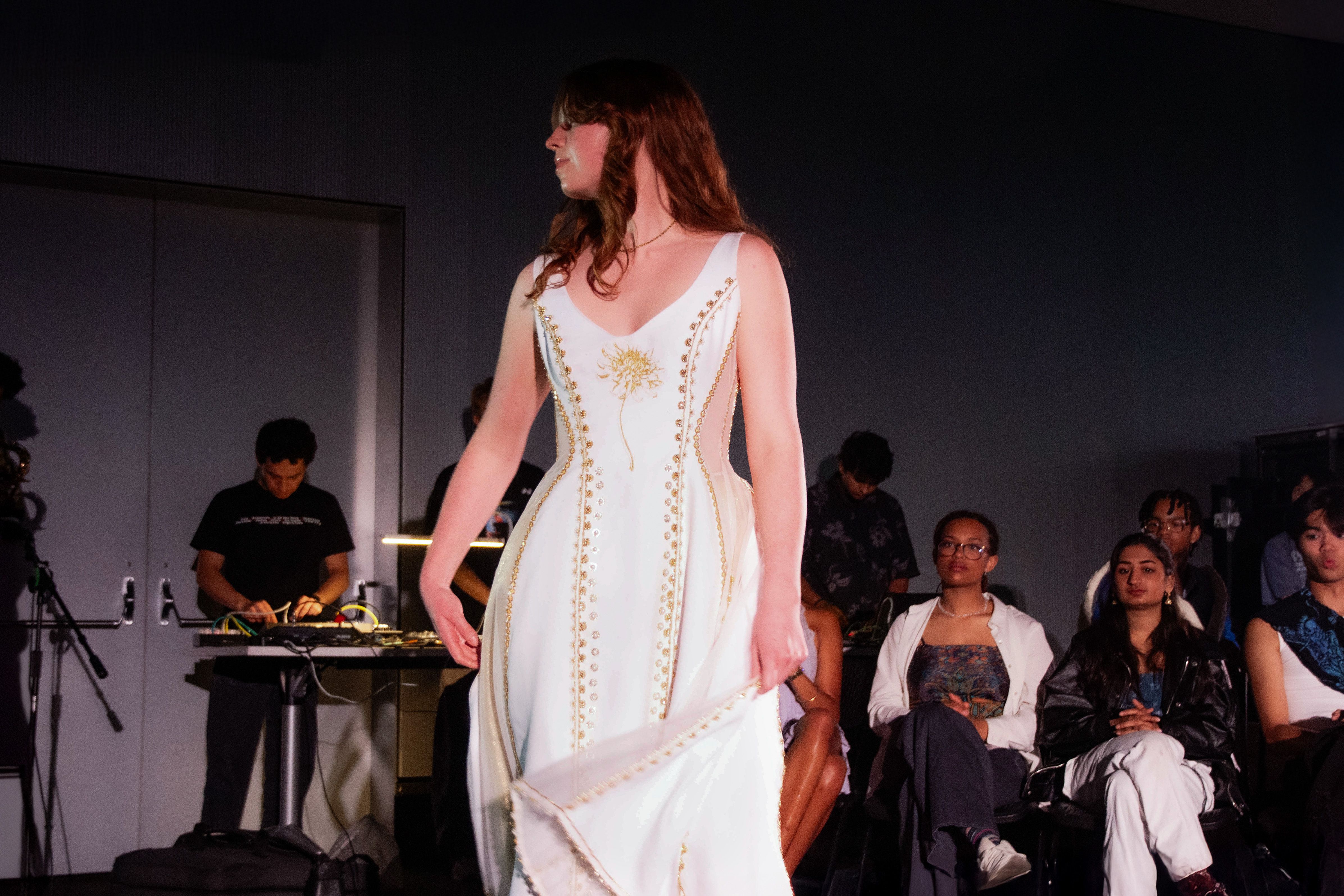 Model walks in a white dress featuring gold embroidery and a sunburst motif on the bodice. The structured gown has a sweeping hemline and is framed by seated spectators.